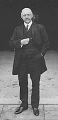 A standing man in three-piece suit, facing camera. He is about 60 and is bald with a mustache. His left hand is in his pants pocket, and his right hand is in front of his chest, holding his pocket watch.