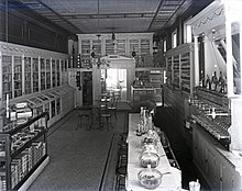 A black and white photograph of the interior of an unidentified drugstore. A large cigar counter and a long bar with seating is featured. The photo was taken by Charles F. Mensing around 1900. The photo is a glass plate negative.