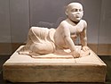 Beige marble statue of a stout young child aged about two years old lying on his left side. The child's head is shaved, his eyes gaze over the viewer's shoulder and his lower body is covered in a draping cloth that hangs limply between his flexed feet. The child supports his torso with his left hand in which he holds an unidentifiable object, he also holds a small bird in his right hand. The sculpture rests on a heavy socle inscribed with barely visible letters spanning the upper part of the socle vertically.