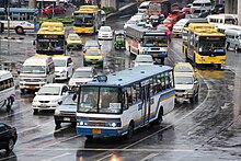 A partial view of a large traffic circle, with many vehicles on the road, including buses, cars, minibuses, brightly painted taxis and a tuk-tuk