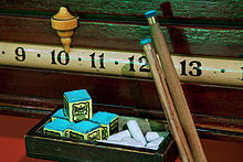 Close-up view of a horizontal scoring band showing the numerals 9, 10, 11, 12, and 13, with a ceramic pointer resting above the 10, two cue tips resting against the 12, and a box of chalk sitting on a shelf beneath