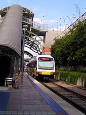 An escalator descends from the street to an island platform station with a white and yellow train present along a landscaped track.