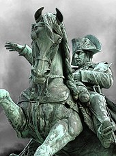 Photo of a grey and phosphorous-coloured equestrian statue. Napoleon is seated on the horse, which is rearing up, he looks forward with his right hand raised and pointing forward; his left hand holds the reins.