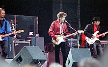 Dylan and members of his band perform onstage. Dylan, wearing a red shirt and black pants, plays an electric guitar and sings.