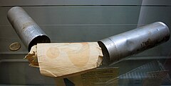 Two metal canisters resting on a glass shelf with a roll of papers, on which a question mark is visible, in between them. A two-euro coin is positioned to the left to provide a scale.