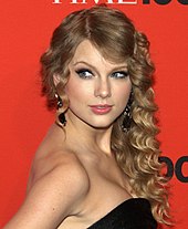 Taylor Swift stands in a Time press area. She has curled hair and is wearing a black, strapless dress.