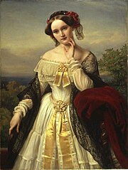 A three-quarter length portrait of a young white woman in the open air. She wears a shawl over an elaborate long-sleeved dress that exposes her shoulders and has a hat on over her centrally parted dark hair.