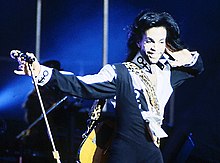 Prince, in a frock and jacket, smiles with a hand to his left ear.