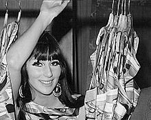 A black and white photograph of a young, dark-haired woman. She's raising her right arm, looking to the right and smiling. She is behind a curtain.