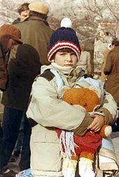 A child wearing winter clothes and holding a large teddy bear looks at the camera, with other similarly dressed adults busy in the background