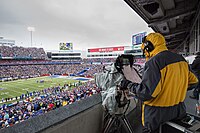 A cameraman with a yellow jacket operates a television camera behind a crowd at a football stadium