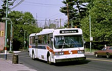 On the roof of the white trackless trolley, two poles rise up to contact overhead wires. A red stripe wraps around the front quarter of the vehicle continuing as a blue stripe on the side.