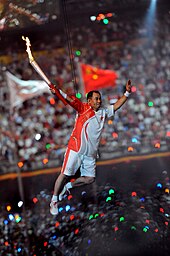 An Asian man in red and white athletic shirt and shorts, and wearing athletic shoes, is suspended by wires in the air while holding a lit torch. In the background, a large crowd in a stadium can be seen, as well as two blurred flags.