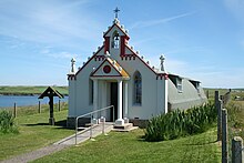 A small chapel sits in green fields under a blue sky. A body of water lies to the left. The front of the building is painted red and white and is decorated with colonnades and a small bell tower. By contrast, the main part of the building is painted grey and has a curved exterior reminiscent of a Nissen hut.