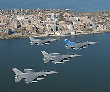 Four jets flying right in formation over water. In the foreground are buildings erected on a narrow piece of land, with water on both sides