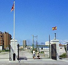 Two women standing at a closed line of gates, with the British and Spanish flags flying on separate poles on either side.