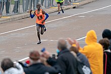 Black man wearing blue arm sleeves, navy shorts and orange sleeveless shirt printed number 2, running down paved street as fans behind barricade cheer him on in foreground