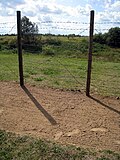 A narrow strip of bare raked soil, on which some footsteps are visible; behind the strip there is a simple barbed-wire fence supported by round wooden posts. Open landscape is visible through the fence.
