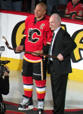 A man in full hockey uniform and another man in a dark suit hold a gold hockey stick together as they look toward an unseen photographer.