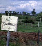 Behind a faded white sign saying "HALT ZONENGRENZE", there is a derelict barbed-wire fence supported by leaning wooden posts; some distance behind that, there are two newer-looking barbed-wire fences supported by concrete posts. Another fence and a guard tower are visible in the distance.