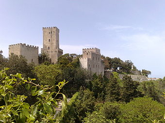 A castle with a tall narrow tower and walls topped by battlements stretches along the edge of a cliff covered in trees and palm trees