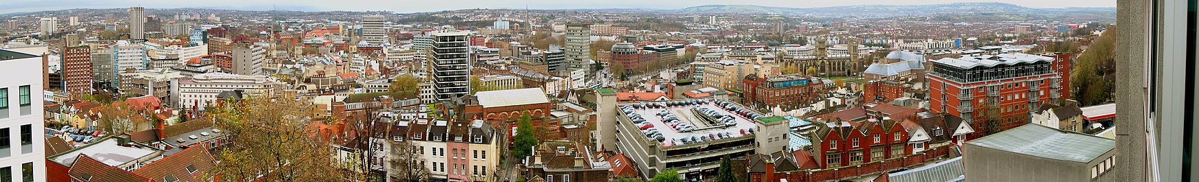 A panoramic view looking over a cityscape of office blocks, old buildings, church spires and a multi-story car park. In the distance are hills.