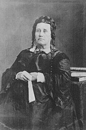 Portrait of an unsmiling, middle-aged woman in a voluminous dress. Her hair is piled on the back of her head, with ringlets near her ears. She holds a fan in her hands.