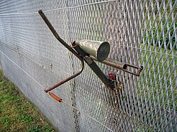 Horn-shaped device mounted on the side of a metal fence, with trigger wires attached to it and running parallel to the fence into the foreground and background.