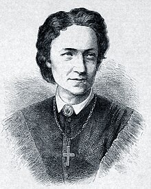 Una mujer de mediana edad con cabello negro hasta los hombros mirando a su izquierda vestida con una camisa de cuello alto y una gran cruz en una cadena