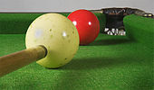 A close-up view of a pockmarked white ball to the front and left of a red snooker ball which is itself next to a corner pocket in top right-hand corner of the image. The tip of a cue stick is visible in bottom right-hand corner of the image, about to strike the white ball.