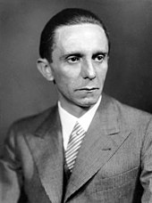 A black and white photo of a man wearing a suit and tie. His body is facing to the left while his head is turned towards the right.