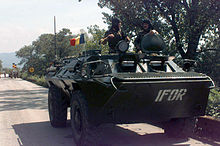 A green tank with white letters spelling "IFOR" drives on a paved road lined with trees.