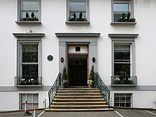 A flight of stone steps leads from an asphalt car park up to the main entrance of a white two-story building. The ground floor has two sash windows, the first floor has three shorter sash windows. Two more windows are visible at basement level. The decorative stonework around the doors and windows is painted grey.