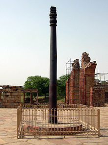 A pillar, slightly fluted, with some ornamentation at its top. It is black, slightly weathered to a dark brown near the base. It is around 7 meters (23 feet) tall. It stands upon a raised circular base of stone, and is surrounded by a short, square fence.