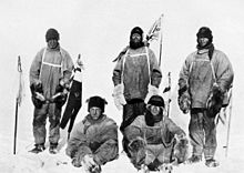 Five men in heavy polar clothing. All look unhappy. The standing men are carrying flagstaffs and a Union flag flies from a mast in the background.
