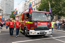 Two firefighters in red "LFB PRIDE" tshirts walk in a parade alongside a fire engine. The fire engine is decorated with at least five rainbow flags.