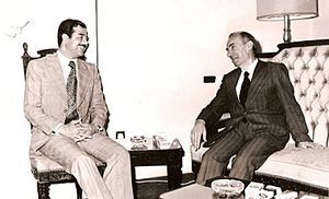 Saddam Hussein and Michel Aflaq seated, talking. Both wear suits.