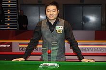 Ding smiling at the camera stands behind a snooker table with his German Masters Trophy.