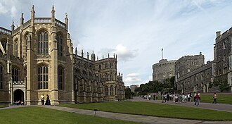 A photograph of a large Gothic chapel on the left, with tall thin windows. On the right is a line of stone buildings, pointing towards a circular tower in the middle of the picture. In the centre are two paths surrounded by grass, with a number of people walking around.
