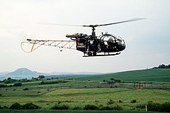 Green-painted helicopter with "Bundesgrenzschutz" on the side flies parallel to a border fence with a gate in it, behind which are two East German soldiers and a canvas-sided truck.