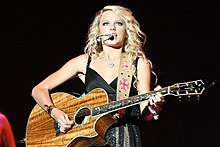Taylor Swift singing on a microphone and playing a guitar. She is wearing a black dress