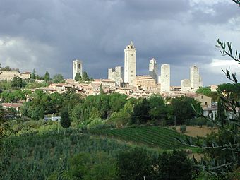 View of a small town on a hilltop surrounded by trees and vineyards. There are eight tall square towers rising from among the densely packed houses.