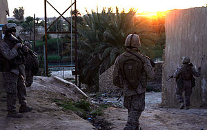Tres infantes de marina patrullan a través de una ciudad iraquí cerca de un río mientras se pone el sol.