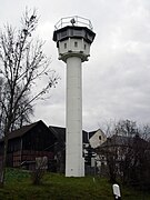 A tall white tower standing near houses and a barn; its base is formed by a slim round column, at the top of which is an octagonal cabin with top-hung windows facing into all directions; there is a searchlight on top of the tower's flat roof, and a railing around the roof's outer edge.