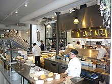 A broad, bright kitchen space with mostly silver and gray tones and warm yellow lights and several chefs at various stations preparing food