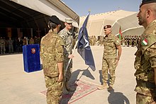 A general hands a NATO flag from a soldier on the left to one on the right.