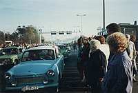 East German Trabant cars driving between dense crowds of people. Metal gantries over the road and a watchtower are visible in the background.