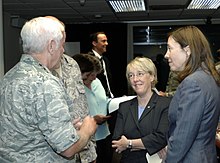 Two adult women talk with an older white-haired man in camouflage inside a dark room.