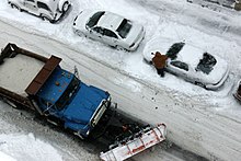 Street seen from above covered in snow with a city truck full of snow and a person who has to remove snow from his or her car