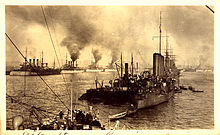 A fleet of warships lined up in a harbour with thick clouds of smoke coming out of their funnels